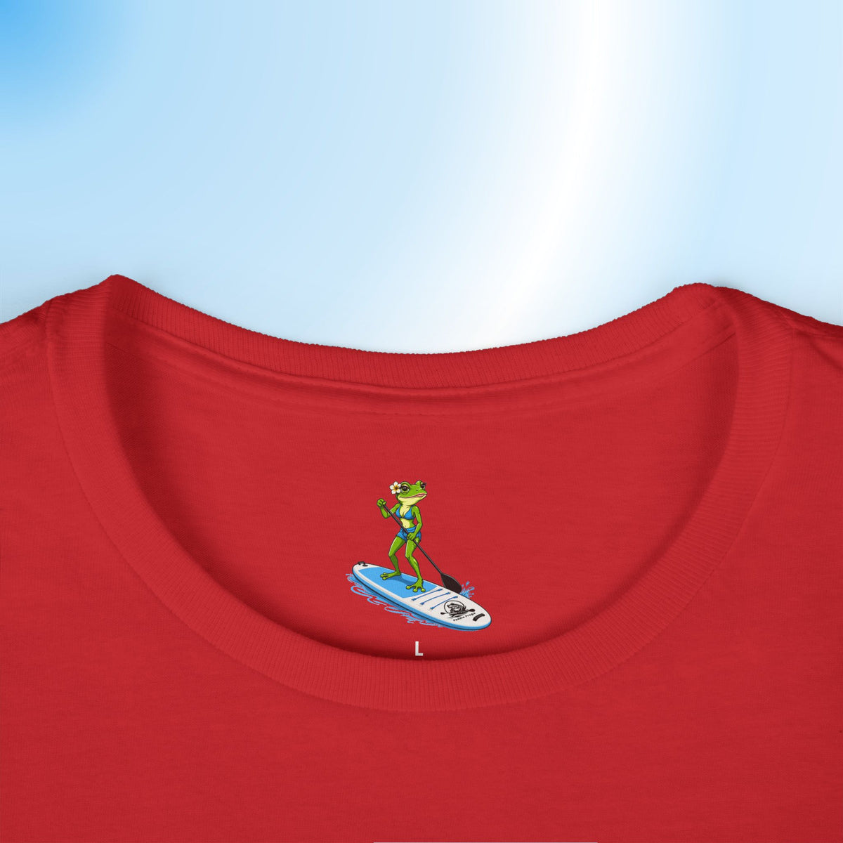 A close-up of a printed neck label on a red ladies crewneck t-shirt showing a small graphic of a green frog standing on a blue and white paddleboard. The t-shirt is displayed against a light blue gradient background.