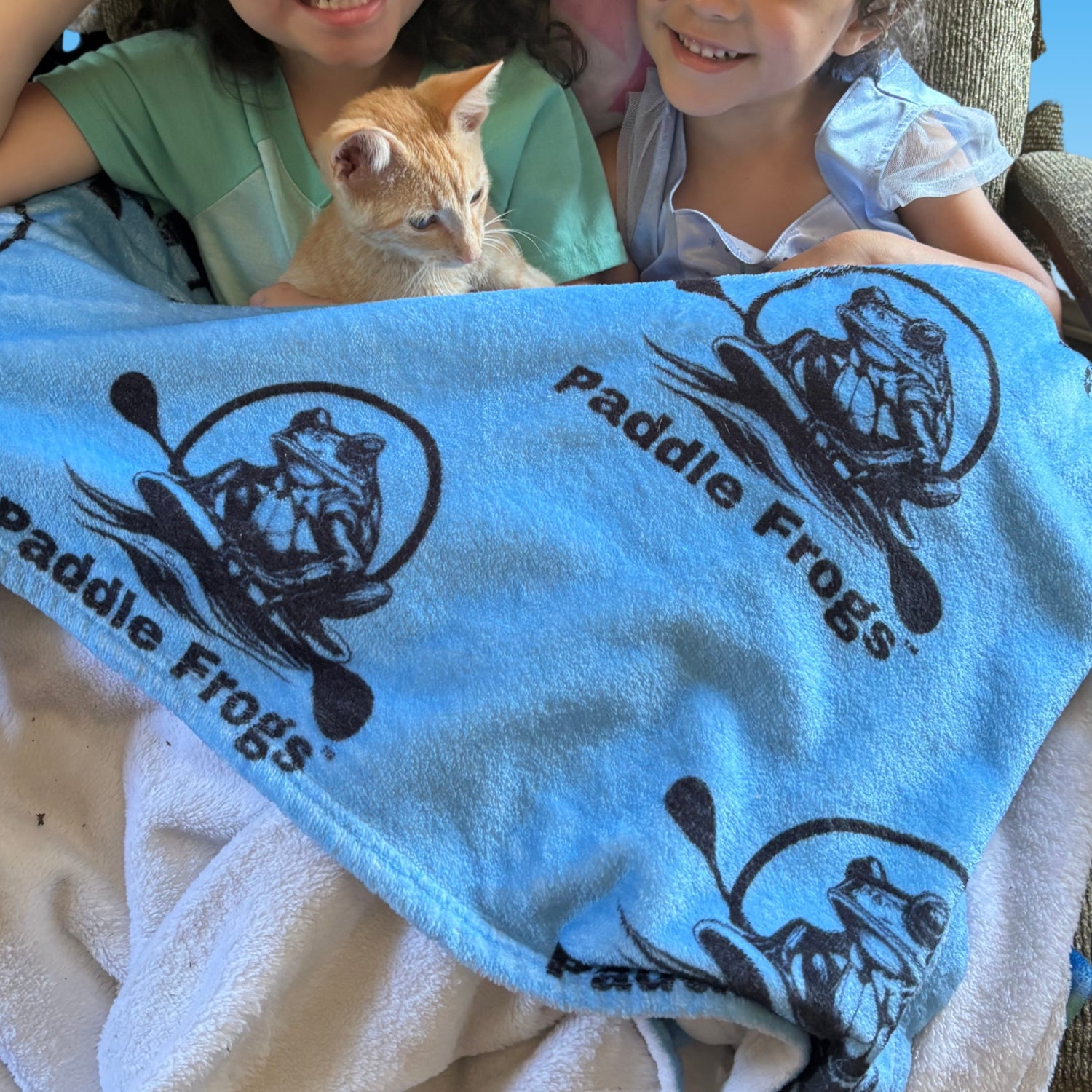 Two children holding a kitten with a blue velveteen blanket featuring 'Paddle Frogs' branding.