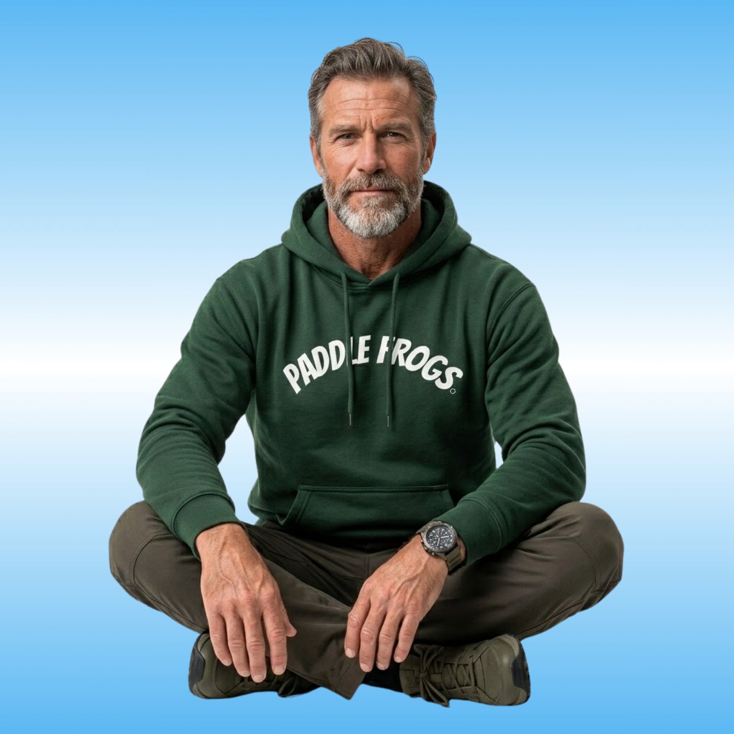 A man with a graying beard sits cross-legged against a light blue gradient background, wearing a dark forest green hoodie with 'PADDLE FROGS' printed retro 90's style in white arched text across the chest.