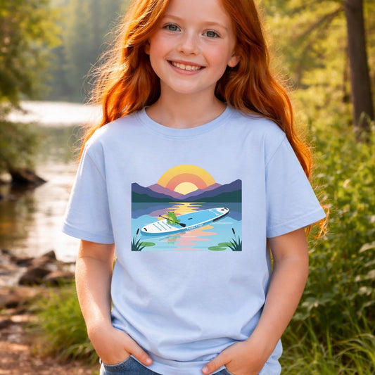 Child wearing a light blue Paddle Frogs t-shirt with a paddleboard design outdoors by the river