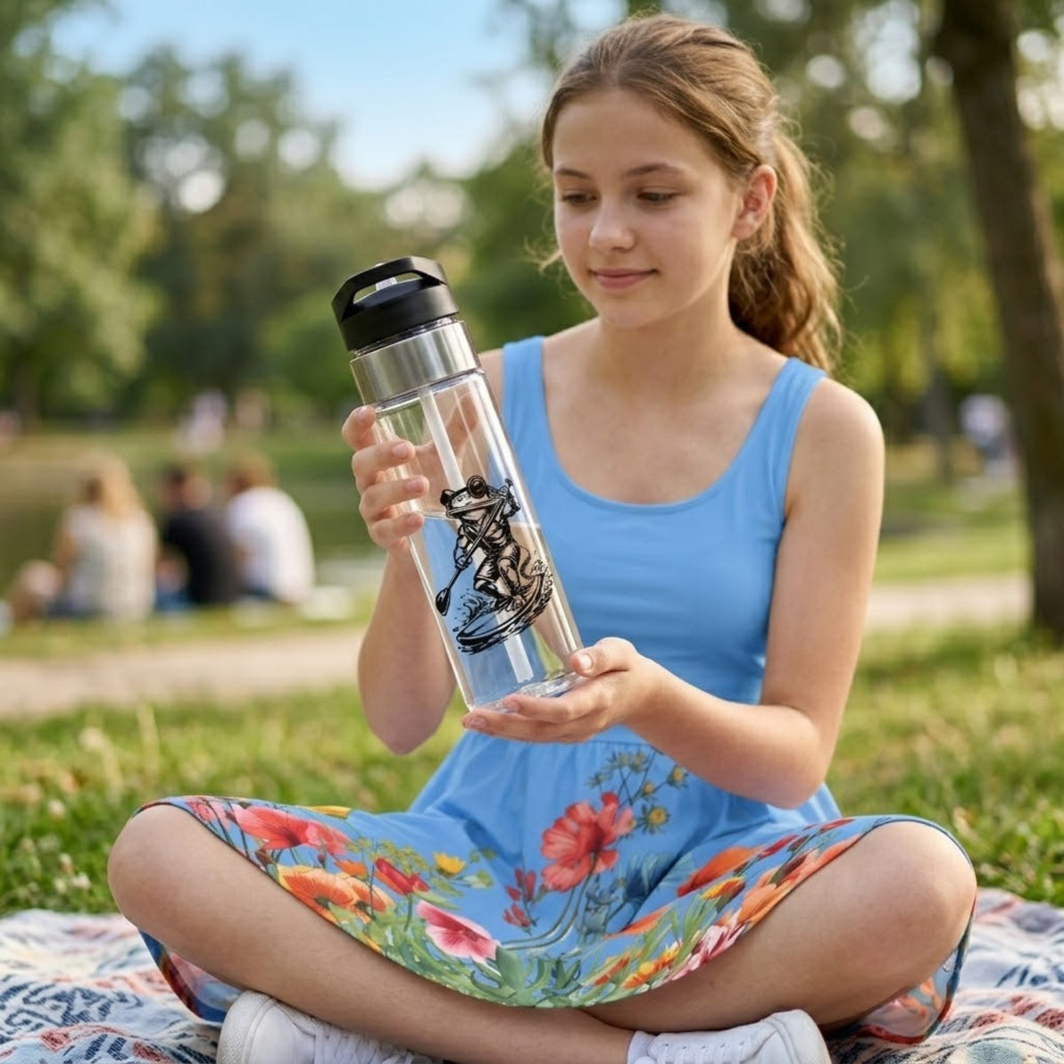 A young girl sitting cross-legged on a blanket in a sunny park, holding a clear plastic water bottle with a black lid and a black graphic of a frog paddleboarding.