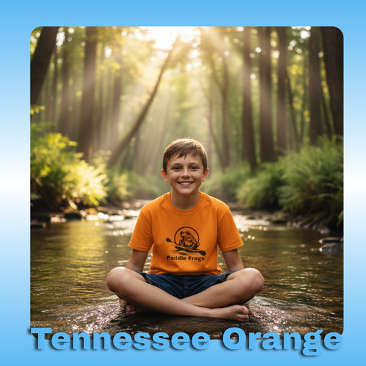 Paddle Frogs Kids T-Shirts | Tennessee Orange with an 11 year old boy in the creek | Child sitting in a stream with sunlight filtering through trees, wearing an orange shirt with 'Paddle Frogs' logo.