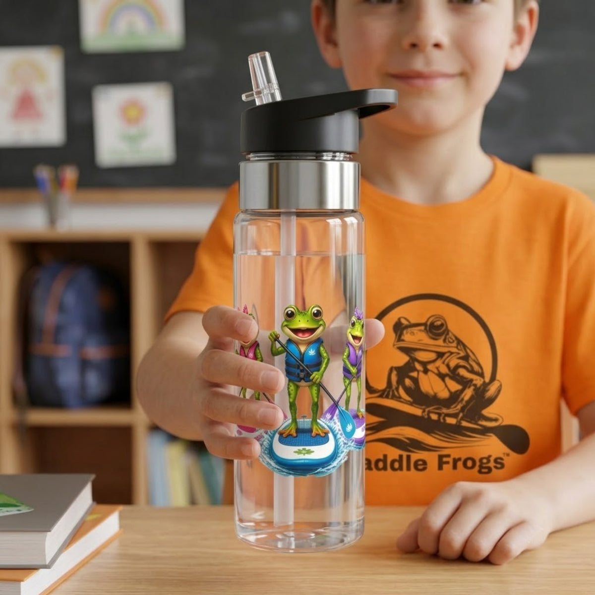 A child in an orange T-shirt holds a clear plastic water bottle with a black lid and straw. The bottle features a colorful graphic of green and purple cartoon frogs paddleboarding.