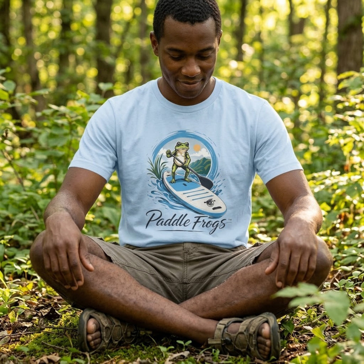 A man meditating outdoors in a forest, wearing a light blue t-shirt with a graphic of a frog on a paddleboard and the text 'Paddle Frogs'.