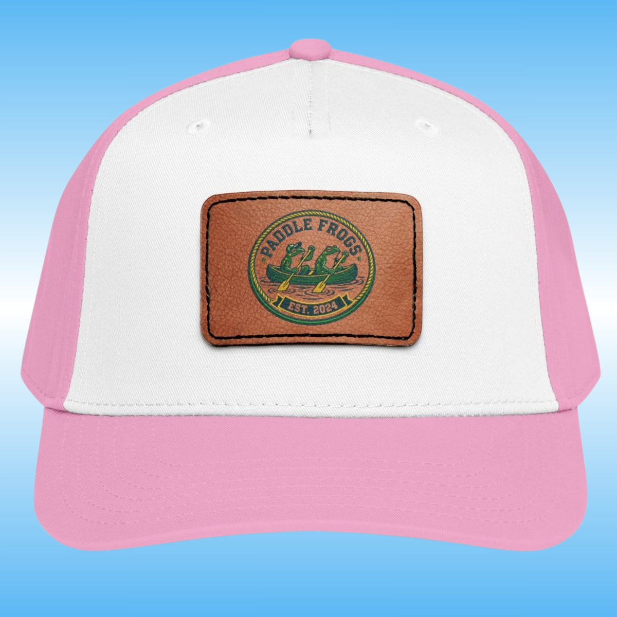 A pink and white trucker-style cap with a brown faux-leather patch on the front featuring the 'Paddle Frogs Est 2024.' canoe logo, set against a blue gradient background.