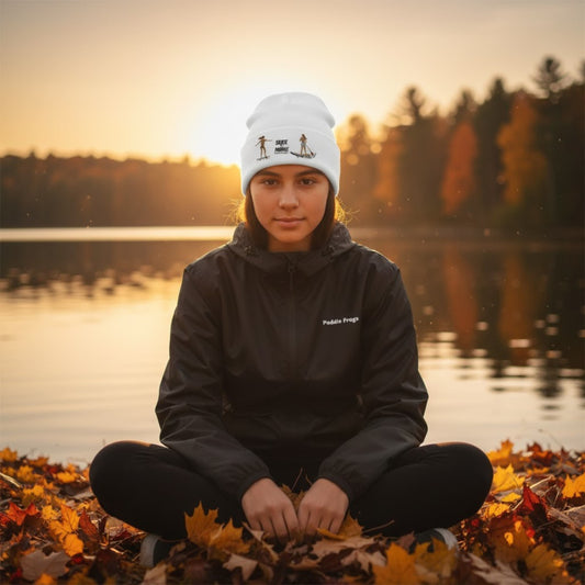 Person sitting on leaves by a lake with a sunset background Tides & Trails Beanie: Life in Balance, Skate & Paddle headwear by Paddle Frogs™ Outdoor Gear sitting crisscross applesauce wearing the packable Ascend Windbreaker - Lightweight Hoodie Jacket