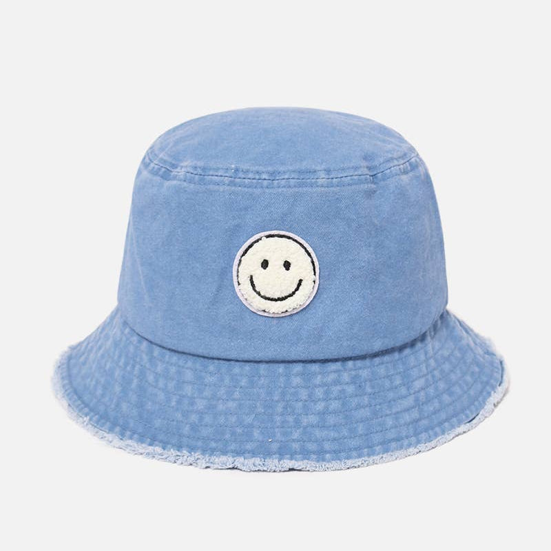 A light blue denim bucket hat with a distressed frayed brim and a circular embroidered smiley face patch on the front, set against a plain light gray background.