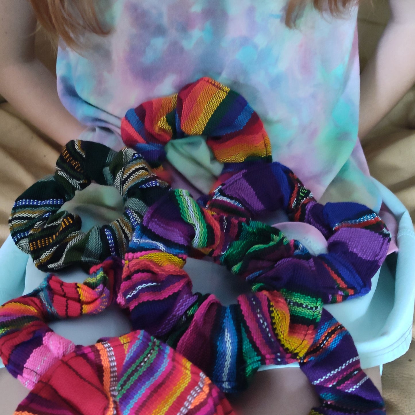 Colorful scrunchies on a white surface with a person wearing a tie-dye shirt in the background.