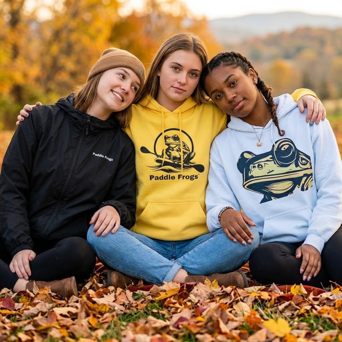three women wearing Paddle Frogs Outdoor Gear clothes yellow original paddleboarding hoodie, Frog Face hoodie in light blue, and Ascend Windbreaker in black. They are sitting crisscross applesauce in autumn season.