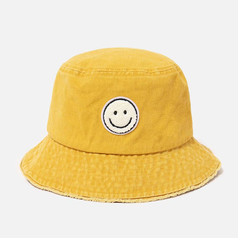 A yellow bucket hat made of textured canvas fabric, featuring a circular embroidered smiley face patch on the front, set against a plain light gray background.