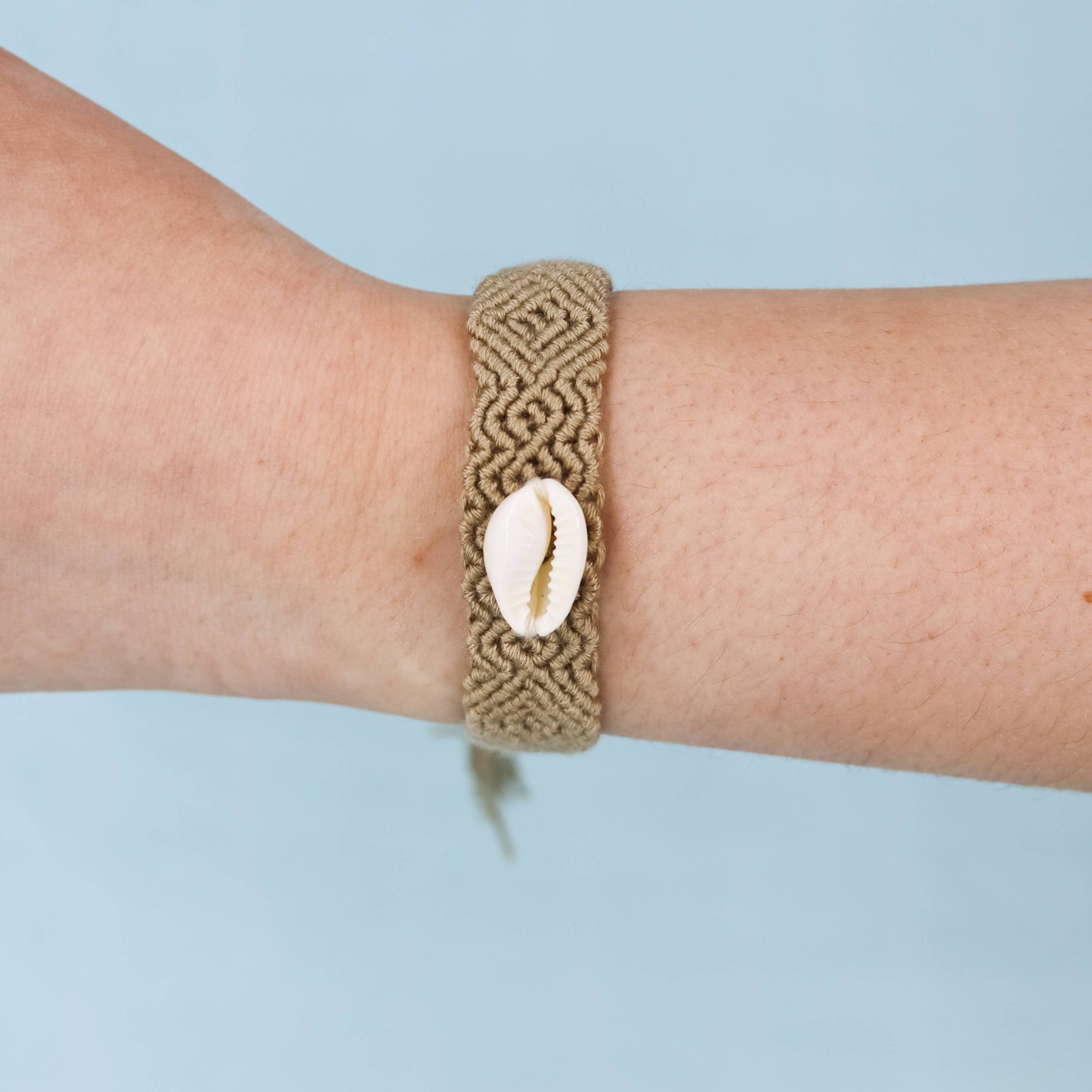 A tan woven macrame bracelet with a white cowrie shell charm, shown worn on a person's wrist against a light blue background.