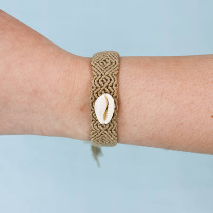 A tan woven macrame bracelet with a white cowrie shell charm, shown worn on a person's wrist against a light blue background.