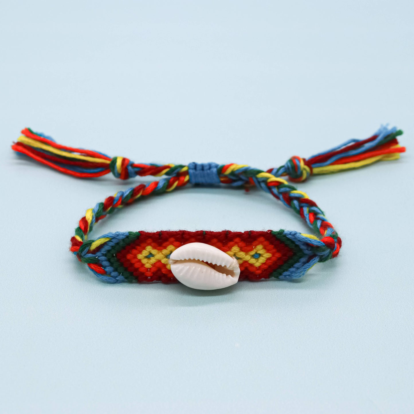 A colorful braided friendship-style bracelet with a central woven geometric pattern in red, yellow, green, and blue, featuring a small white cowrie shell in the center and tassel ties on both ends, shown on a light blue background.