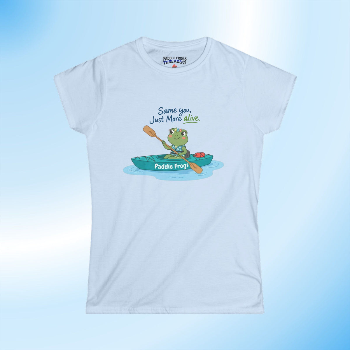 A light blue short-sleeve t-shirt with a cartoon graphic of a green frog paddling in a teal kayak, with the text 'Same you, Just More Alive' and 'Paddle Frogs' on the kayak. The shirt is displayed against a light blue gradient background.