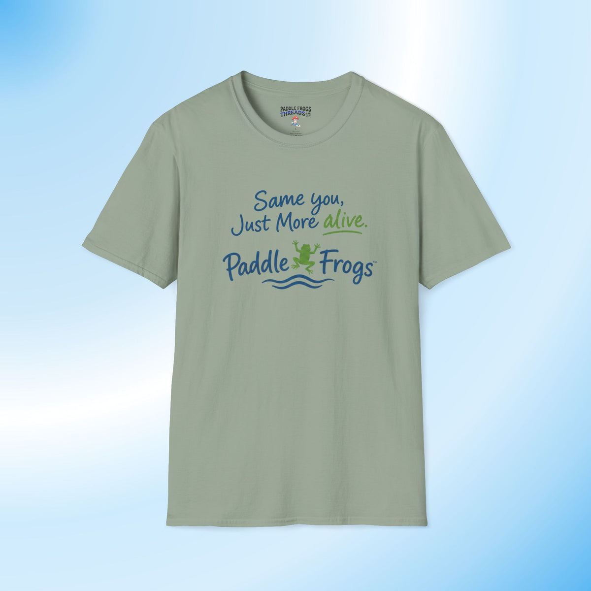 A sage green short-sleeve t-shirt with a graphic on the chest that reads 'Same you, Just More alive. Paddle Frogs' in blue and green lettering with a small frog illustration, displayed against a light blue gradient background.