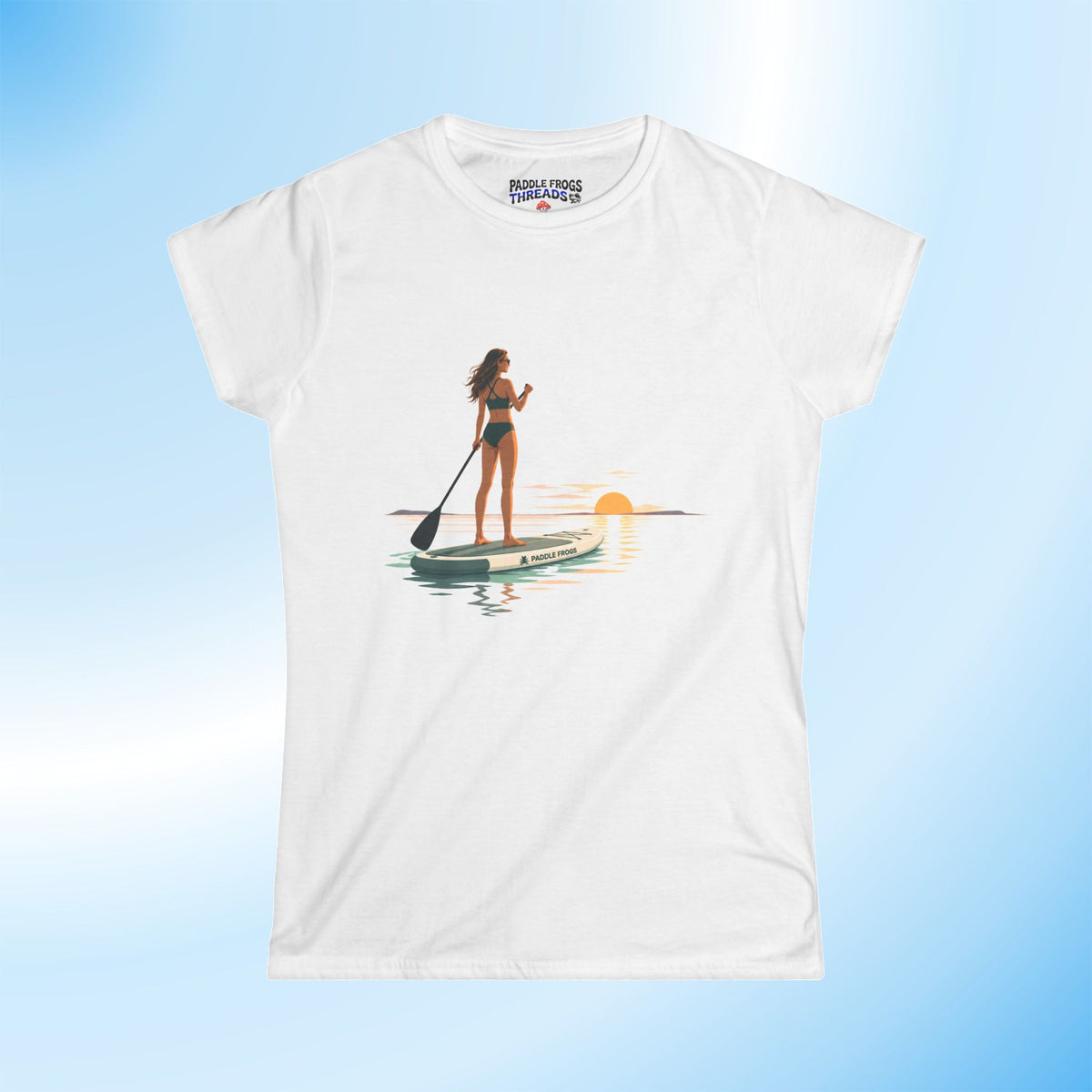 A white short-sleeve crewneck t-shirt featuring a graphic print of a woman stand-up paddleboarding on water at sunset. The shirt is displayed against a light blue gradient background.