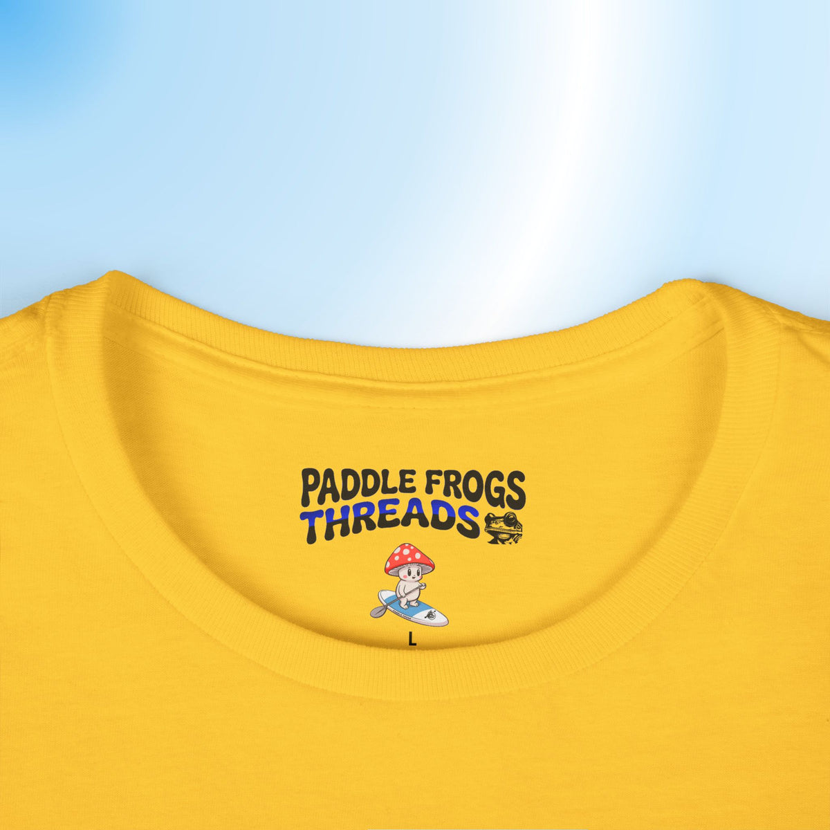 Close-up of a yellow t-shirt neck area featuring a printed graphic that reads 'PADDLE FROGS THREADS' in black and blue lettering, with a small cartoon frog on a surfboard below, set against a light blue gradient background.