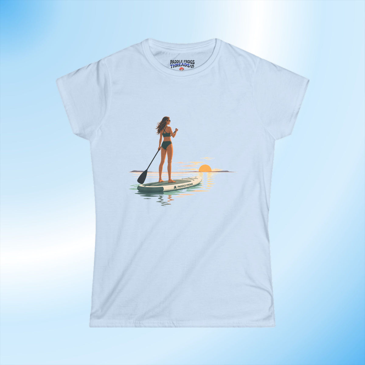A light blue short-sleeve ladies crewneck t-shirt featuring a graphic print of a woman paddleboarding on the water at sunset. The shirt is displayed against a light blue gradient background by Paddle Frogs Outdoor Gear