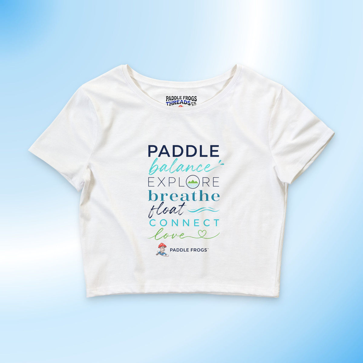 A white short-sleeve crop top t-shirt featuring a colorful graphic text print that reads 'PADDLE balance EXPLORE breathe float CONNECT love' with the Paddle Frogs logo, displayed on a light blue gradient background.