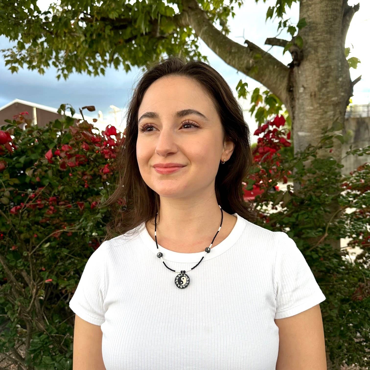 Woman wearing a white shirt with a necklace, standing outdoors with greenery and red flowers in the background.