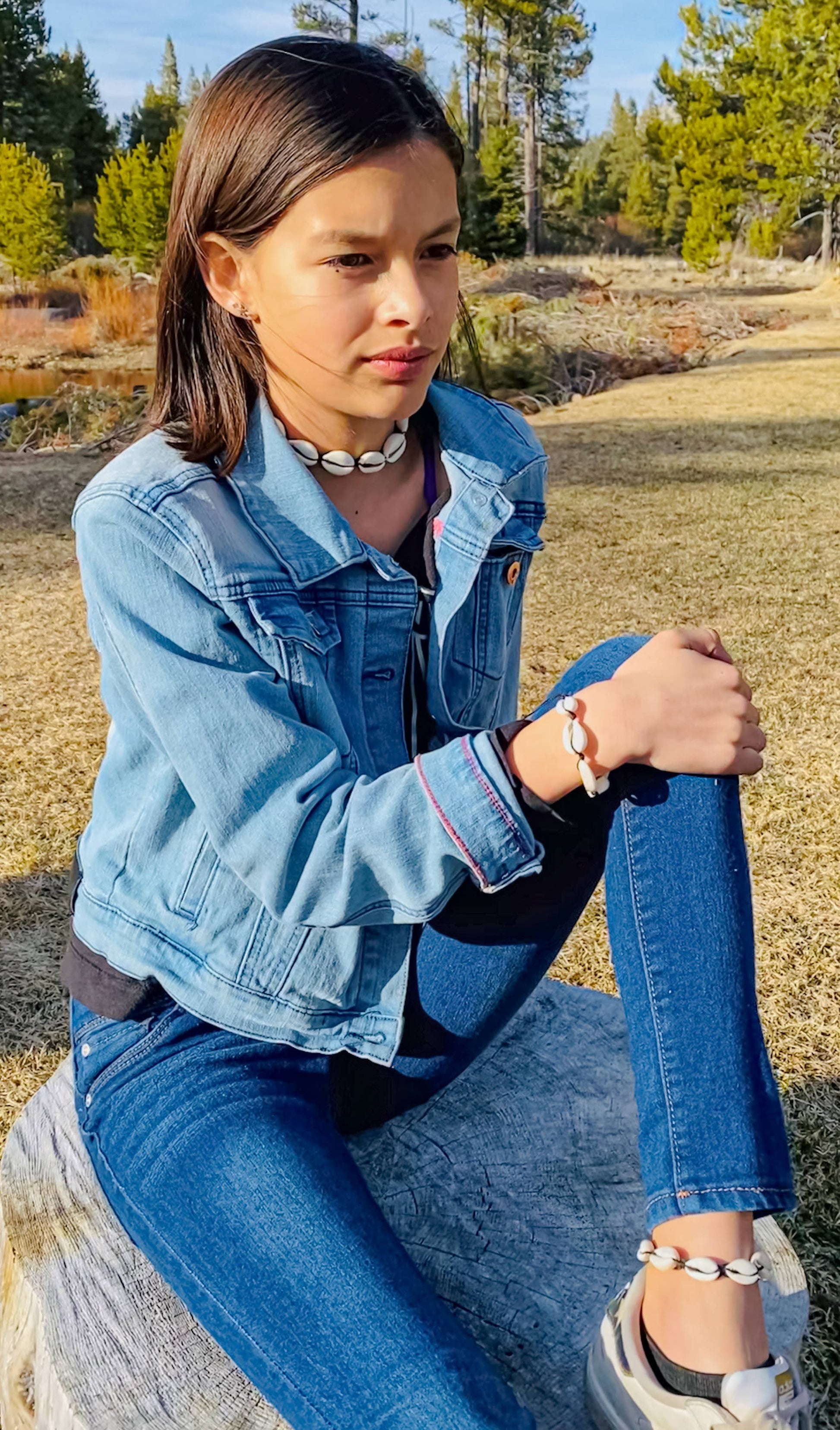Woman wearing a denim jacket and jeans sitting outdoors with trees in the background
