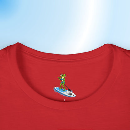 A close-up of a printed neck label on a red ladies crewneck t-shirt showing a small graphic of a green frog standing on a blue and white paddleboard. The t-shirt is displayed against a light blue gradient background.
