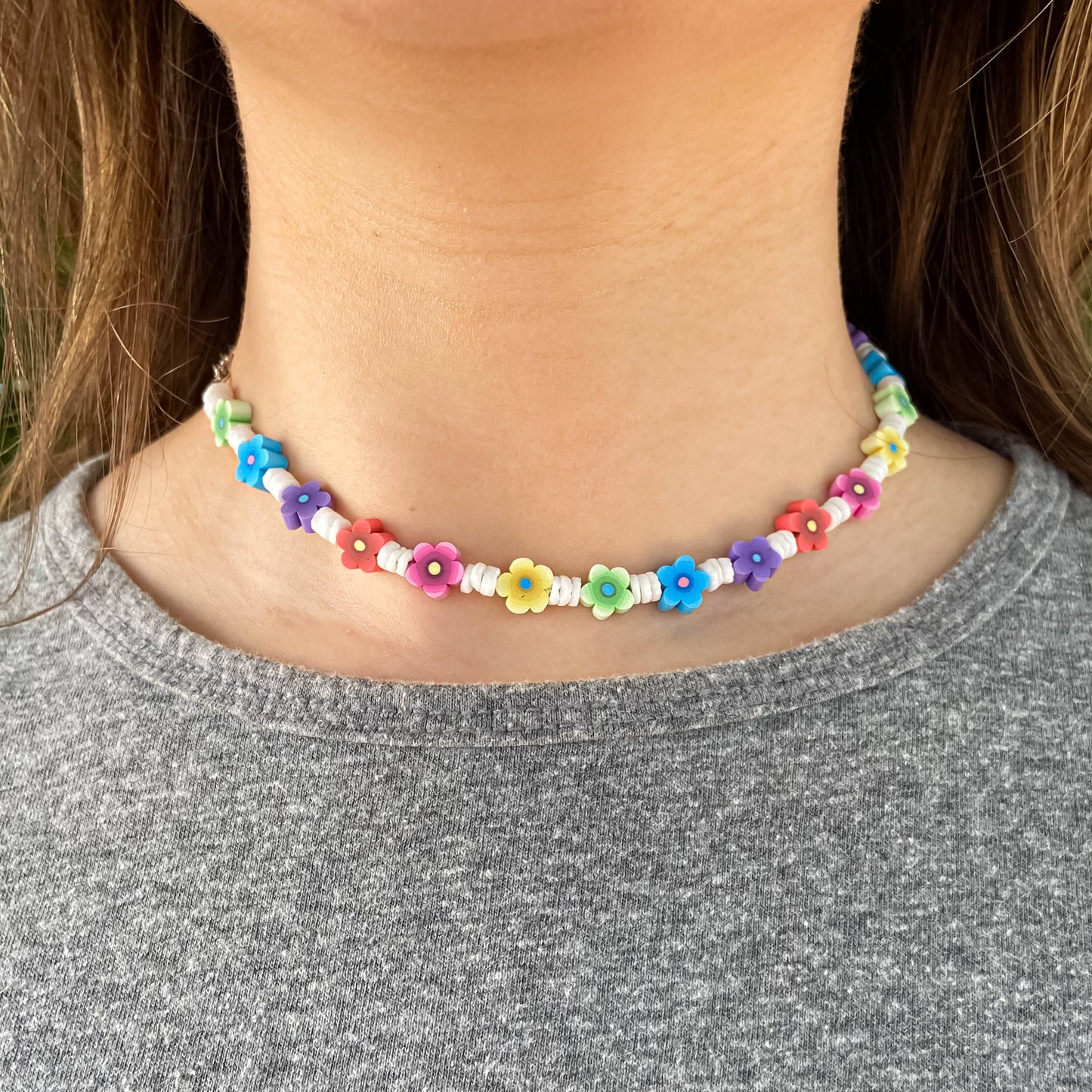 A close-up of a person wearing a beaded choker necklace made of small colorful flower beads and spacer beads, paired with a gray heathered t-shirt.