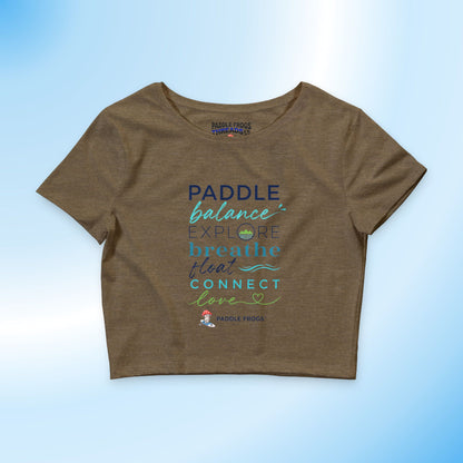 A brown short-sleeve crop top t-shirt featuring a colorful graphic print with the words 'PADDLE balance EXPLORE breathe float CONNECT love' in blue, teal, and green lettering, displayed on a light blue gradient background.