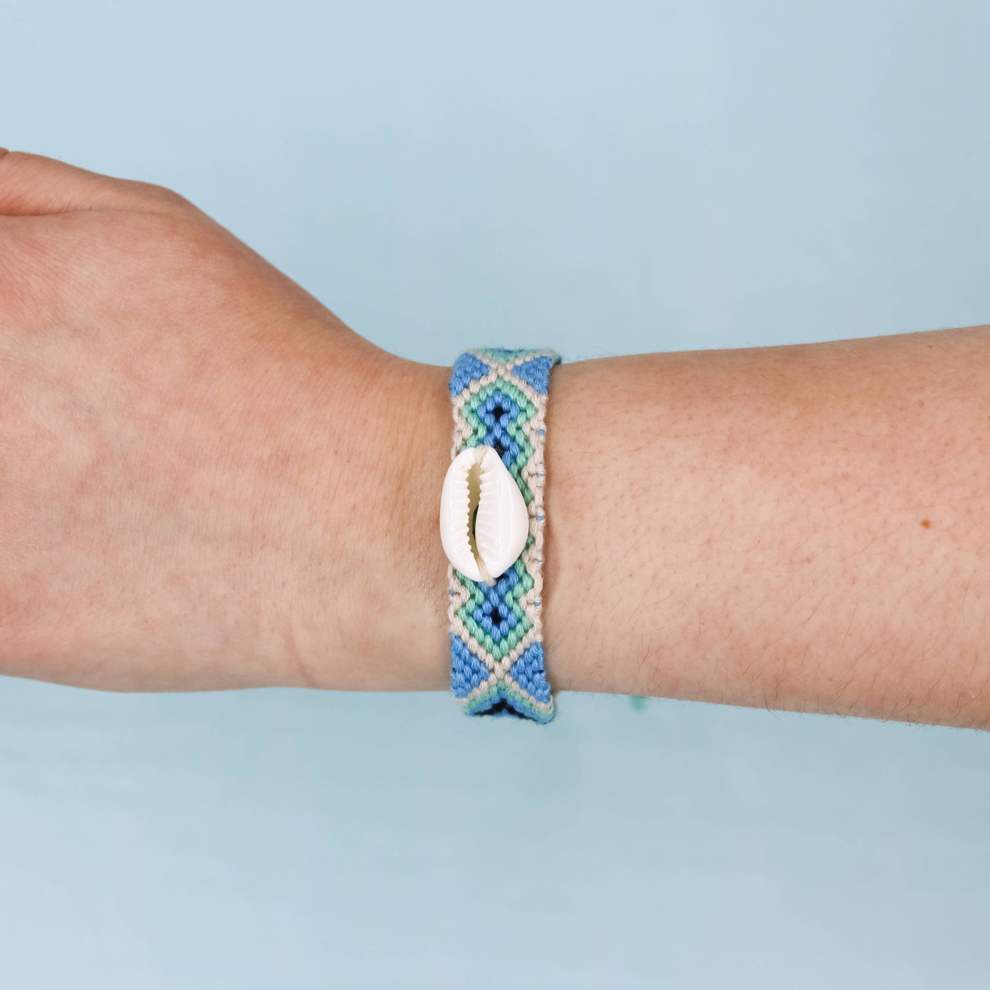 A close-up of a person's wrist wearing a braided blue and green bracelet with a white cowrie shell charm, set against a solid light blue background.