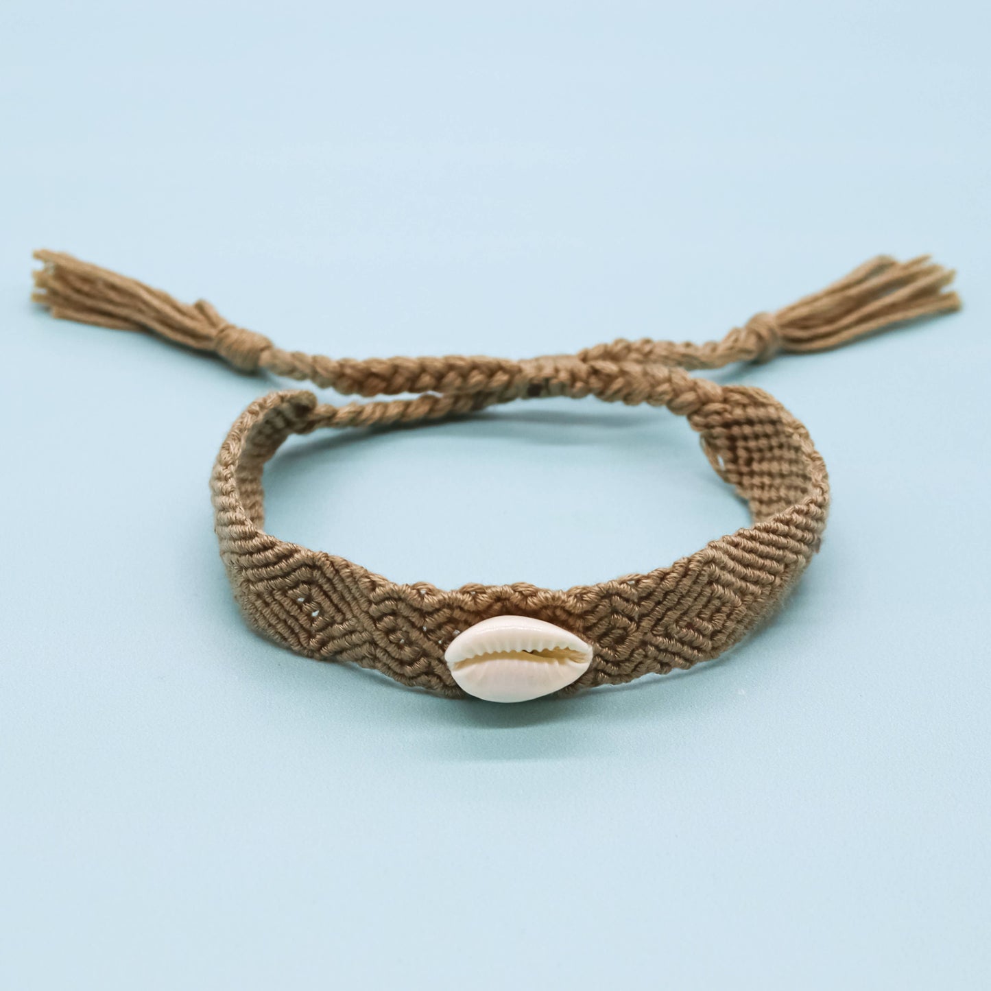 A brown braided macrame bracelet with a small white cowrie shell in the center and adjustable tassel ends, displayed on a light blue background.