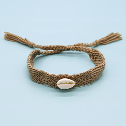 A brown braided macrame bracelet with a small white cowrie shell in the center and adjustable tassel ends, displayed on a light blue background.
