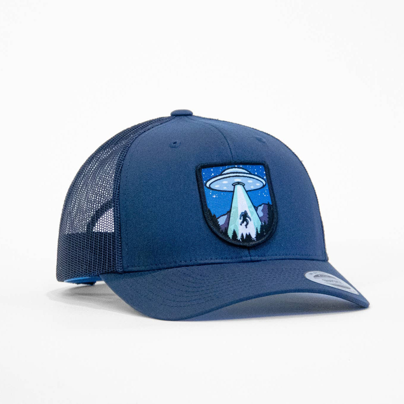 A navy blue trucker cap with a mesh back and a central embroidered patch featuring a UFO sucking up bigfoot and mountain design, shown on a clean white background.