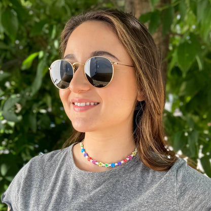 A woman wearing round gold-framed sunglasses with dark lenses and a colorful beaded necklace, photographed outdoors against a blurred background of green leaves.