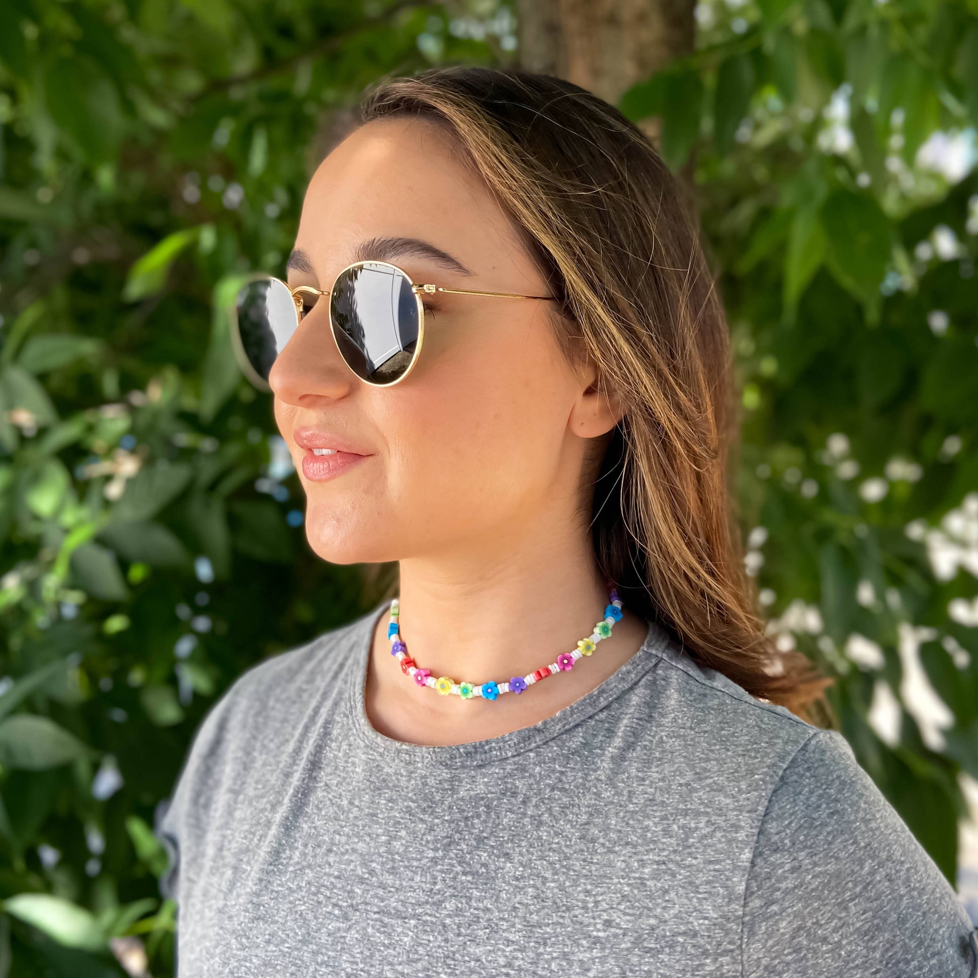 A woman wearing round gold-framed sunglasses and a colorful beaded choker necklace, photographed outdoors against a background of green foliage.