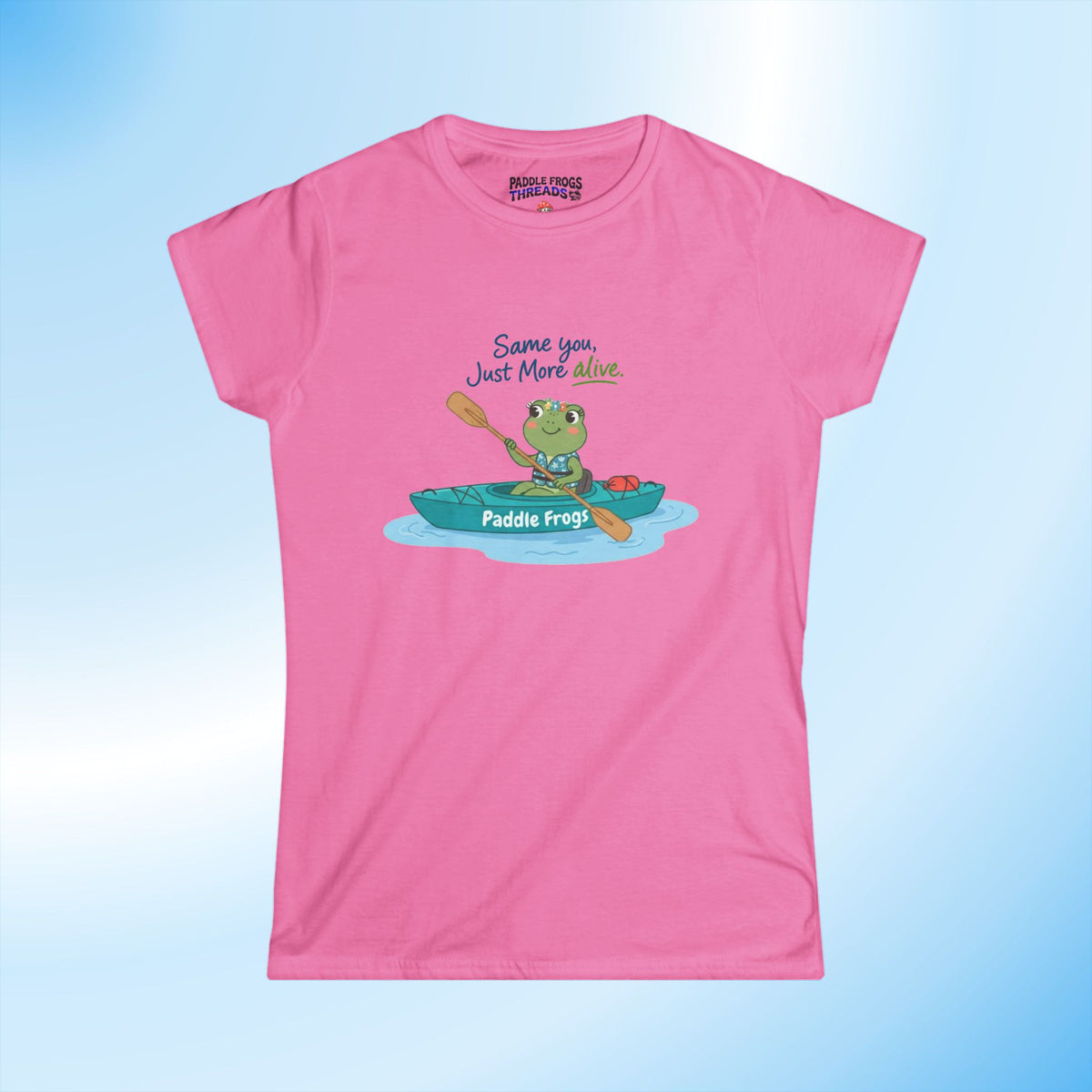 A pink short-sleeve t-shirt with a cartoon graphic of a frog in a teal kayak and the text 'Same you Just More Alive' and 'Paddle Frogs' on the boat. The shirt is displayed flat against a light blue gradient background.
