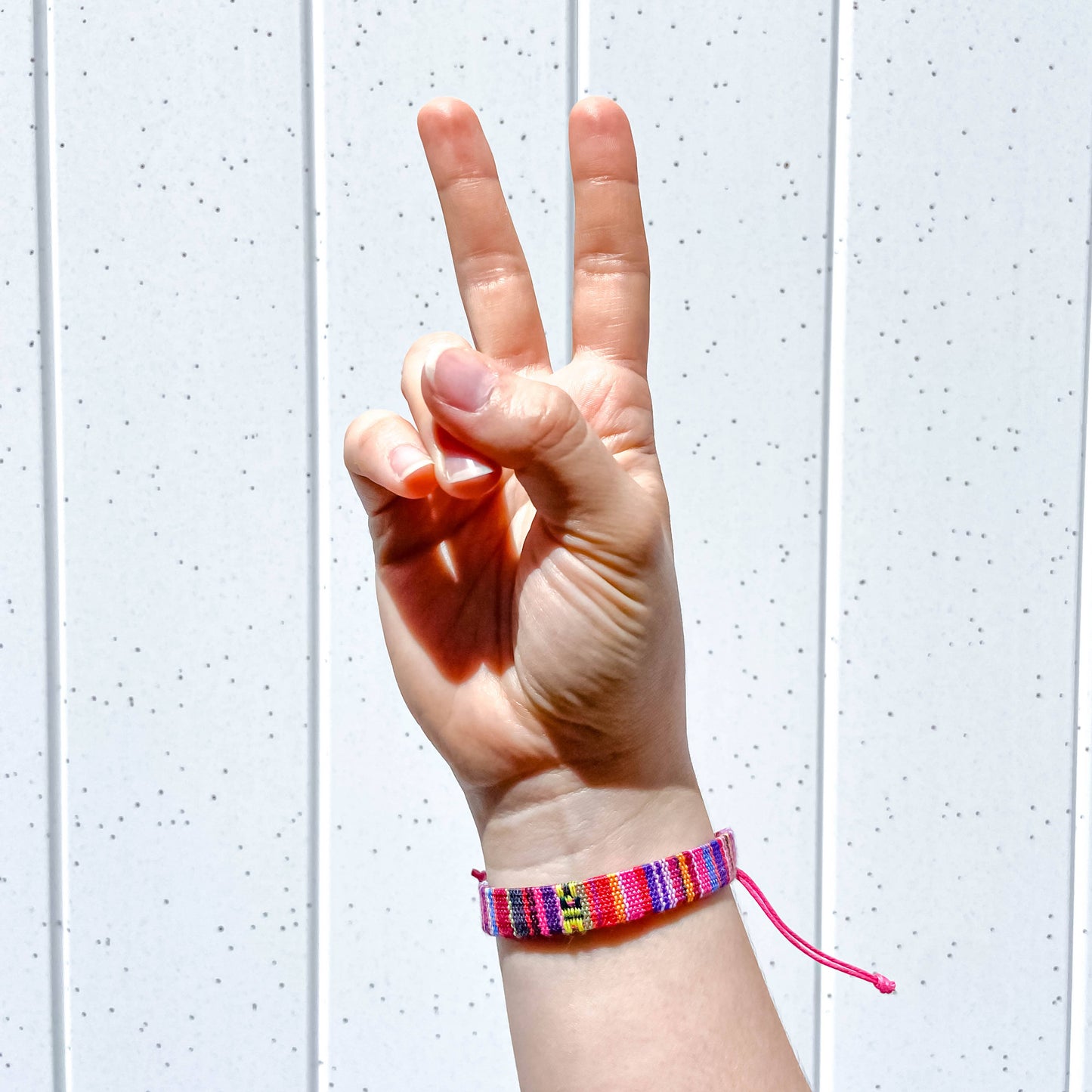 A close-up of a person's hand making a peace sign while wearing a colorful woven bracelet with a small yellow and black flag charm, set against a white paneled background.