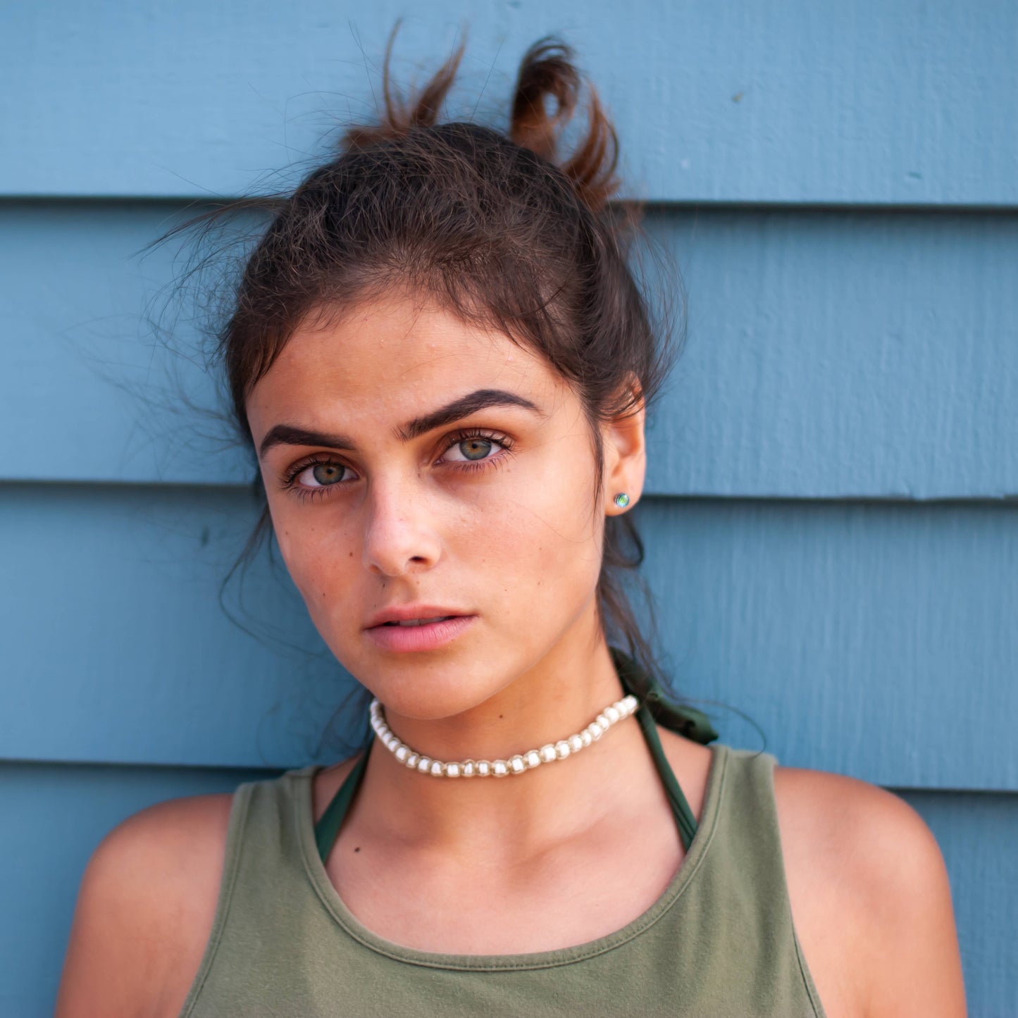Woman with a ponytail wearing a green tank top and a hemp puka shell necklace against a blue wall.