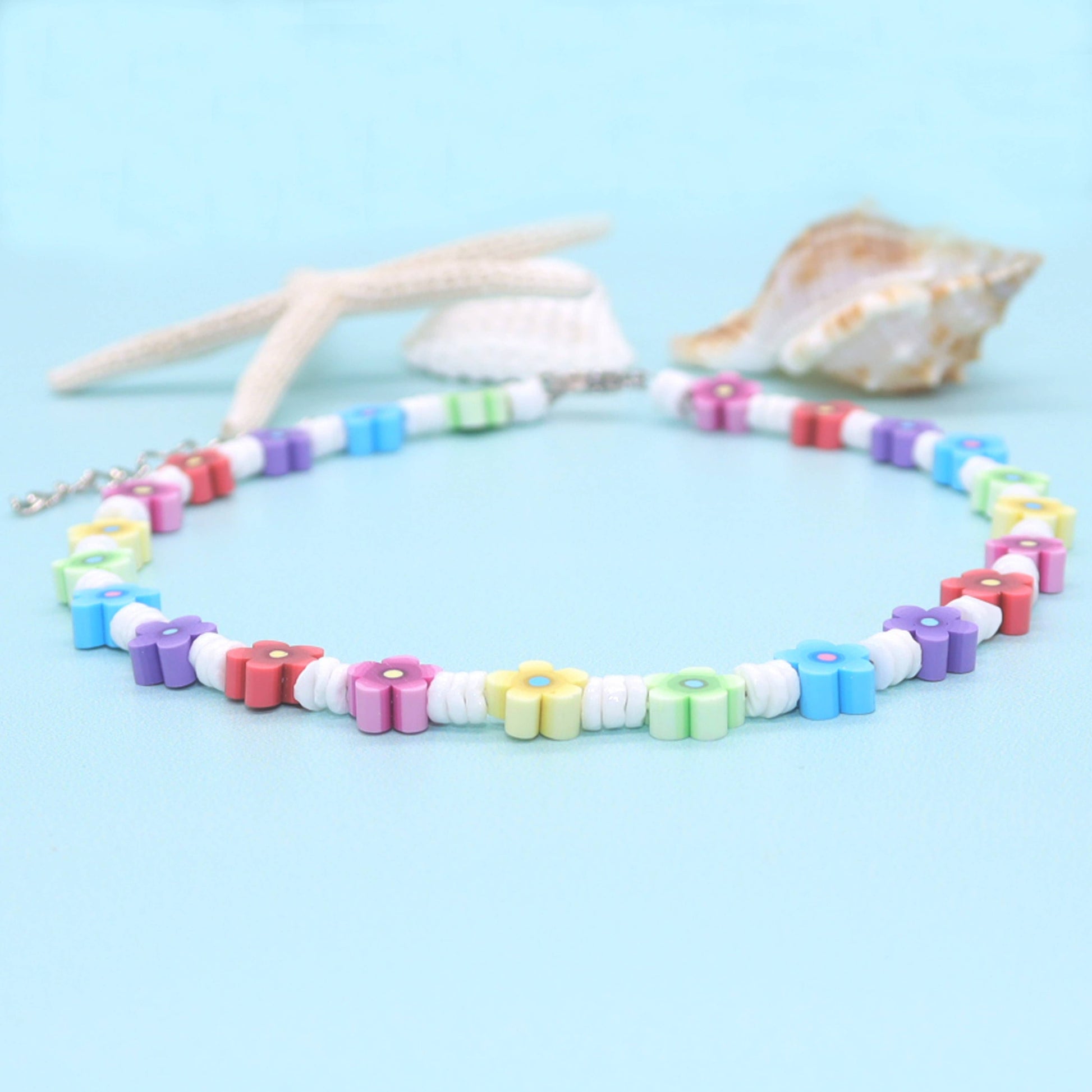 A pastel rainbow flower bead necklace arranged in a circle on a light blue background, styled with a starfish, a small shell, and a larger conch shell.