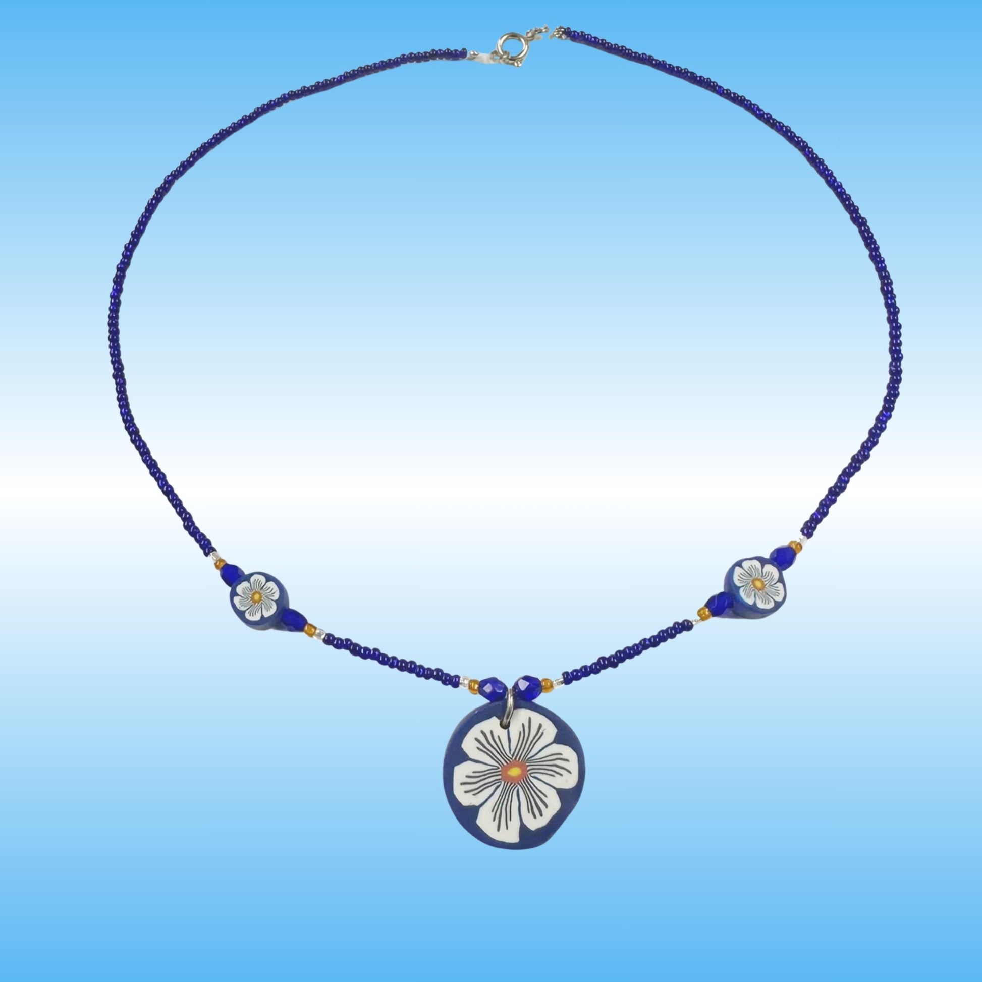 A handmade beaded necklace with a central oval pendant and two smaller floral beads, featuring a dark blue and white design with gold accents, set against a blue gradient background.