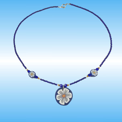A handmade beaded necklace with a central oval pendant and two smaller floral beads, featuring a dark blue and white design with gold accents, set against a blue gradient background.