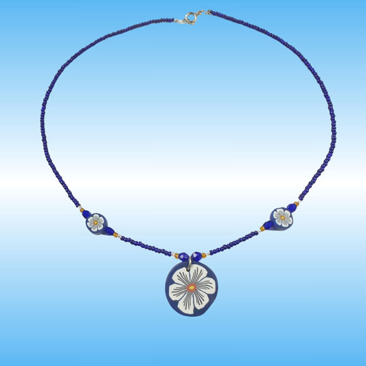 A handmade beaded necklace with a central oval pendant and two smaller floral beads, featuring a dark blue and white design with gold accents, set against a blue gradient background.