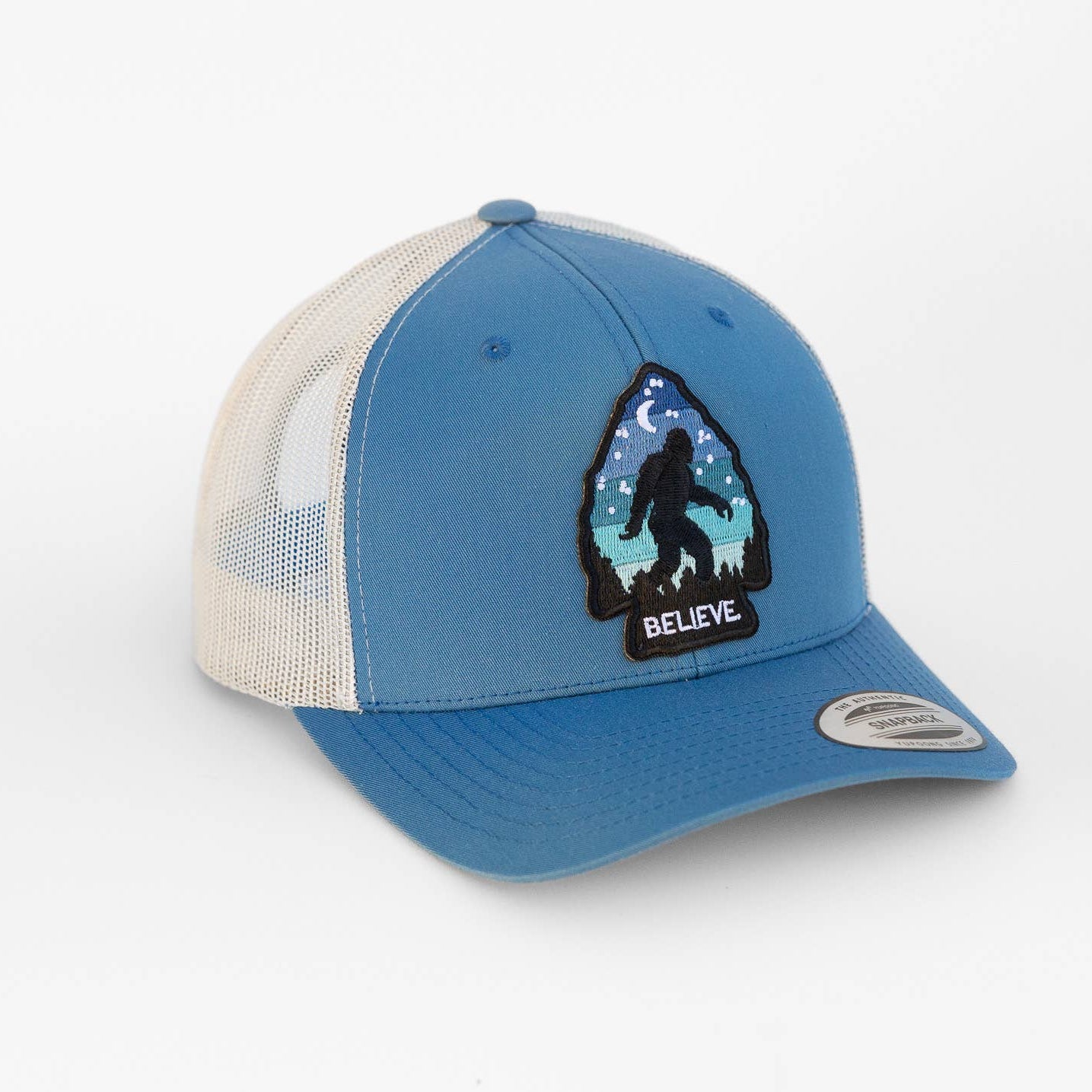 A blue and off-white trucker hat featuring a front patch with a Bigfoot silhouette and the word 'BELIEVE' on an arrowhead.