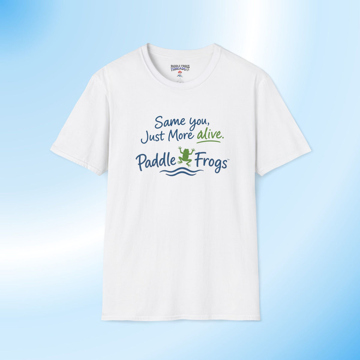 A white short-sleeve crewneck t-shirt with a graphic printed on the chest that reads 'Same you, Just More alive. Paddle Frogs' in blue and green lettering with a small frog illustration, displayed against a light blue gradient background.