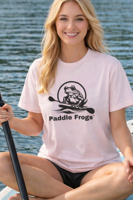 A light pink, short-sleeved cotton t-shirt featuring the Paddle Frogs™ original logo on the center chest. The logo consists of a  frog silhouette holding a paddle, encircled by the brand name. The shirt is shown with a woman sitting crisscross applesauce riding the Divine Escape paddleboard.