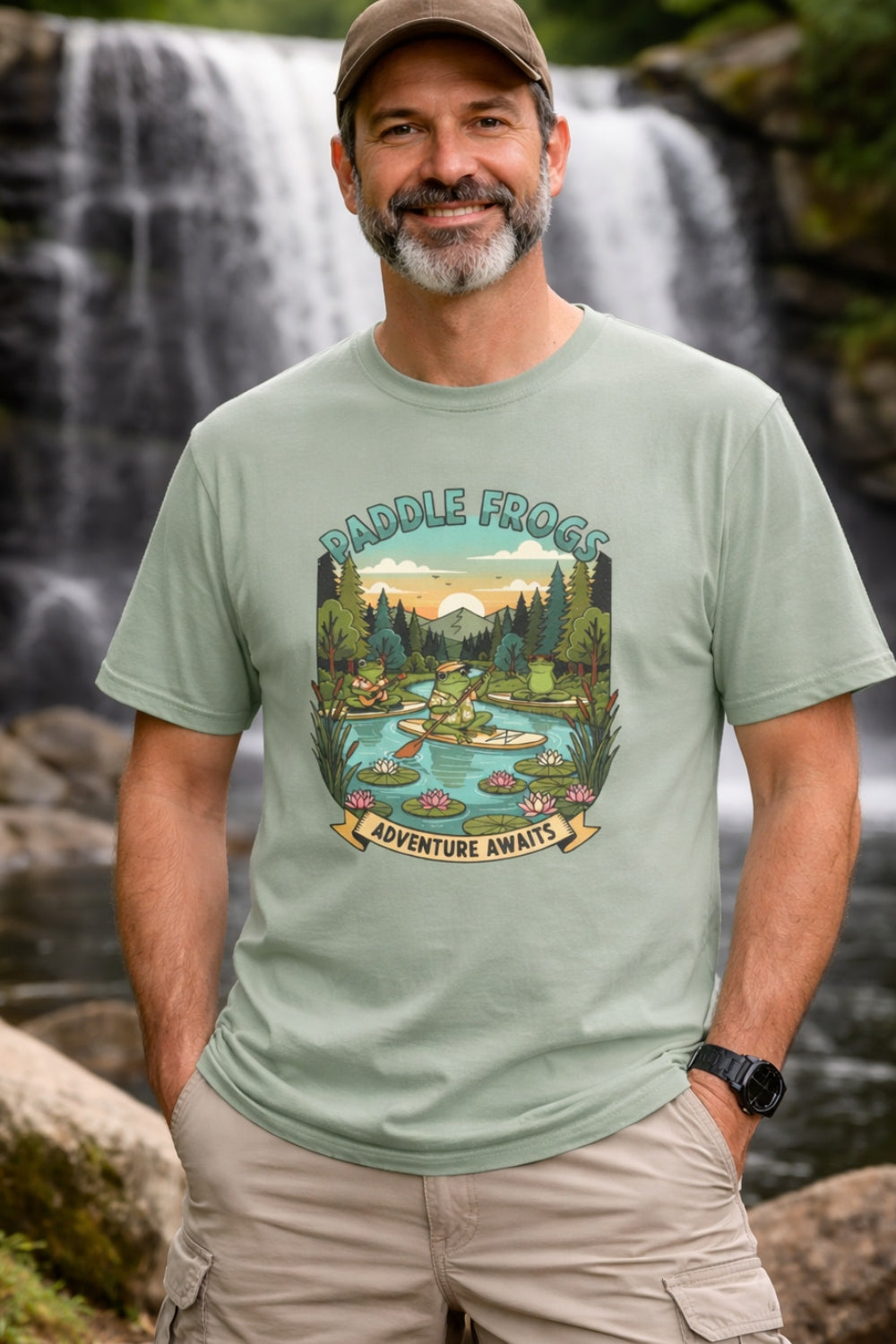 A middle-aged man with a beard and a brown baseball cap smiles in front of a scenic waterfall. He is wearing a light sage green t-shirt featuring a colorful graphic of a frog paddleboarding on a river. The graphic includes the text "PADDLE FROGS" at the top and "ADVENTURE AWAITS" on a banner at the bottom. The man has his hands in the pockets of his tan cargo shorts, and a black watch is visible on his wrist.