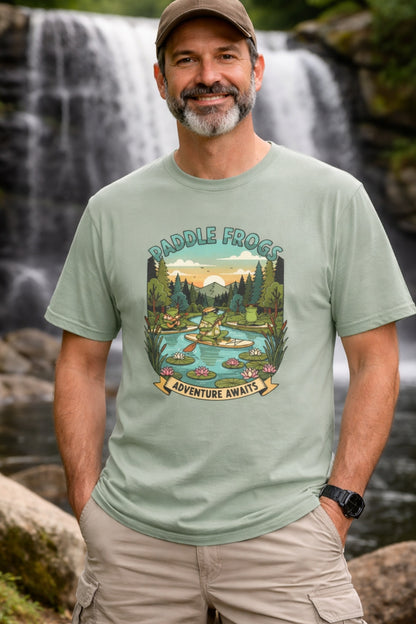 A middle-aged man with a beard and a brown baseball cap smiles in front of a scenic waterfall. He is wearing a light sage green t-shirt featuring a colorful graphic of a frog paddleboarding on a river. The graphic includes the text "PADDLE FROGS" at the top and "ADVENTURE AWAITS" on a banner at the bottom. The man has his hands in the pockets of his tan cargo shorts, and a black watch is visible on his wrist.