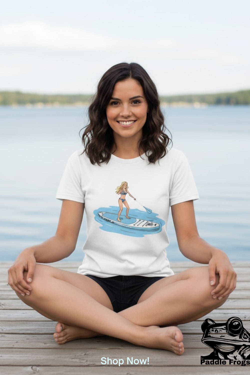 A woman sitting cross-legged on a wooden dock by a lake wearing a white t-shirt with a graphic of a woman on a Divine Escape paddleboard.