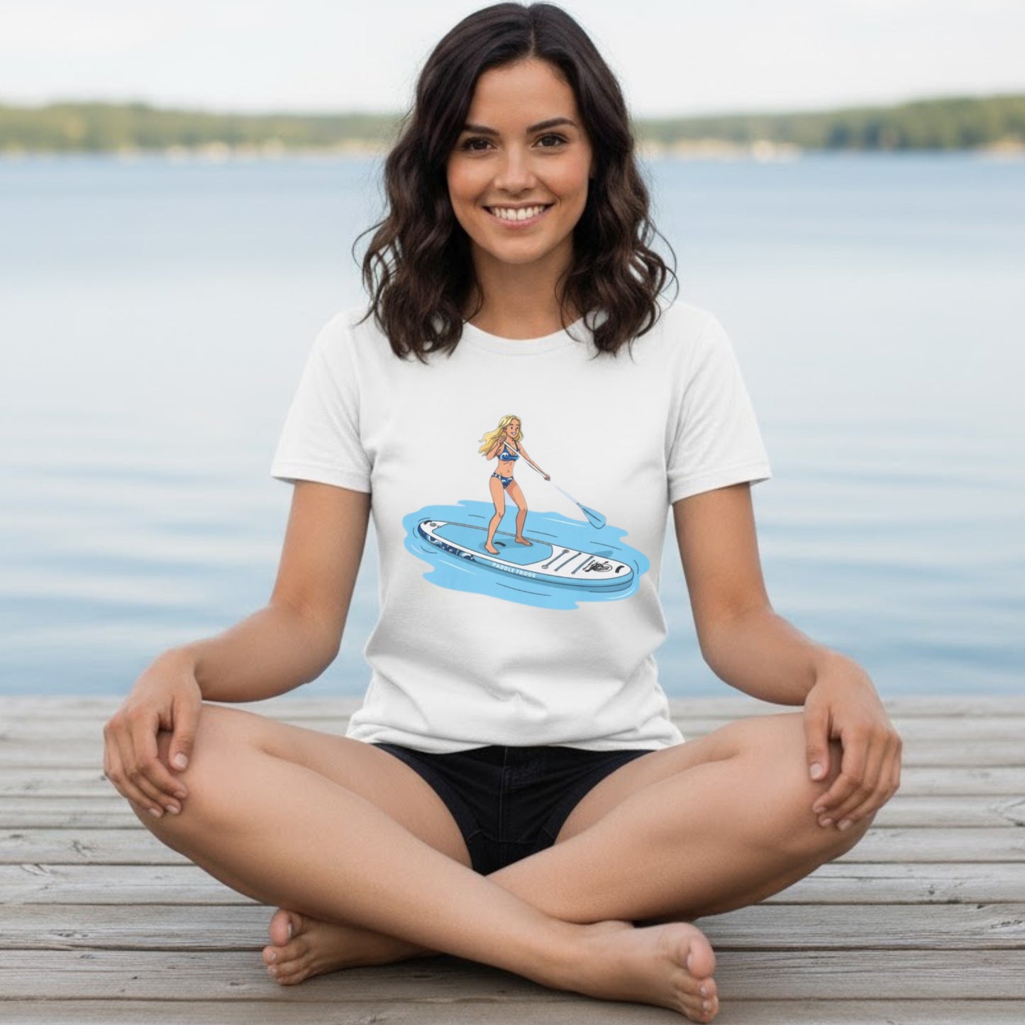 Woman sitting on a dock wearing a white t-shirt with a graphic design of a woman on a paddleboard. Aqua Babe Tee -Divine Escape paddleboard shirt White color sitting crisscross applesauce at Chickamauga lake in Harrison Tennessee