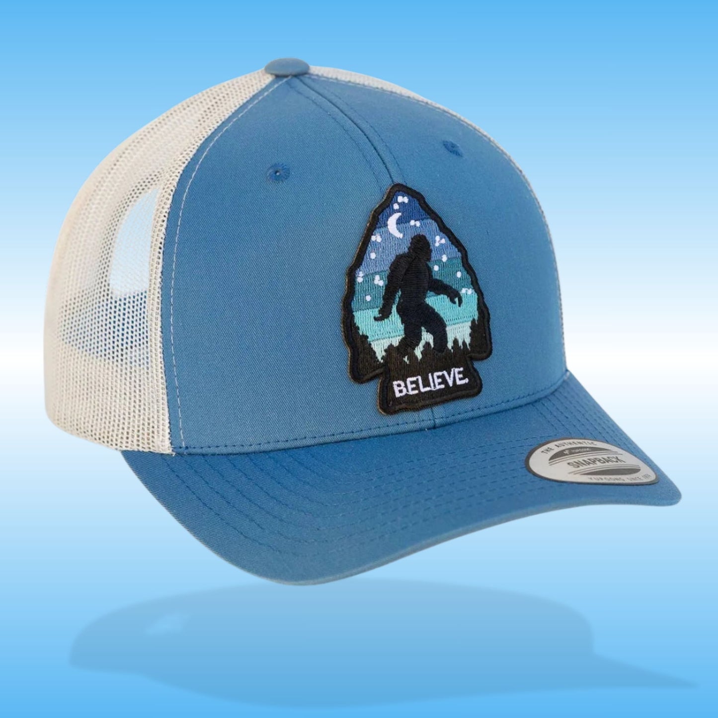 Blue and silver baseball hiking cap with an arrowhead patch saying "Believe" Bigfoot Vintage Trucker Hat on a light blue background.