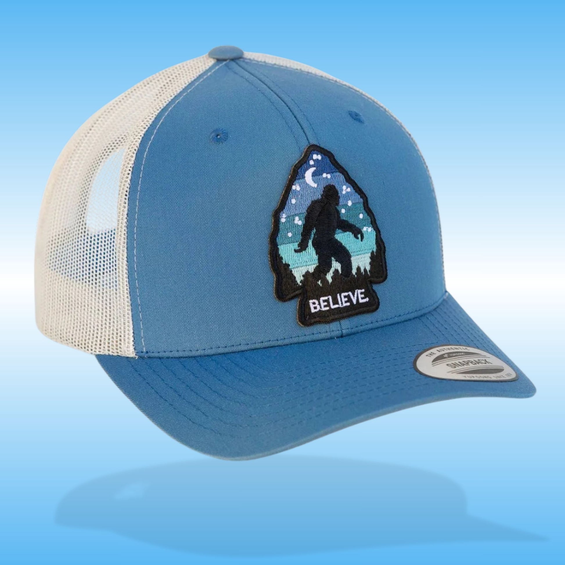 Blue and silver baseball hiking cap with an arrowhead patch saying "Believe" Bigfoot Vintage Trucker Hat on a light blue background.